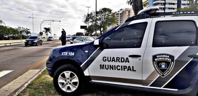 Aspirante da Guarda Municipal analisa quadro digital com vagas em Florianópolis ao fundo